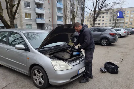 Akuvahetus kortermajade vahelises parklas, tehnik töötab hõbedase auto avatud mootoriruumis, maas tööriistakott, akuklemmid kaetakse pärast pingutamist kontaktmäärdega, mis hoiab ära oksüdeerumise.