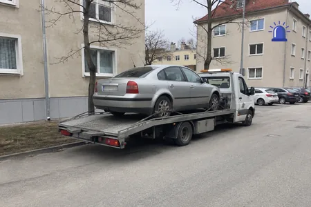 Puksiirabi, hõbedane sõiduauto platvormveokil kortermajade vahelisel tänaval, kaasa sõitev klient istub platvormveoki kabiinis, mitte veetavas autos.